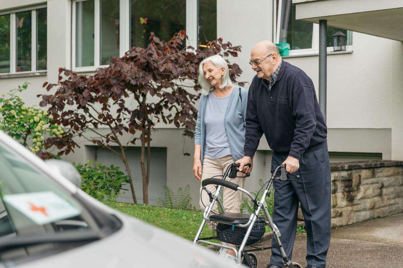 Eine Rotkreuz-Fahrerin begleitet einen Mann am Rollator zum Auto.