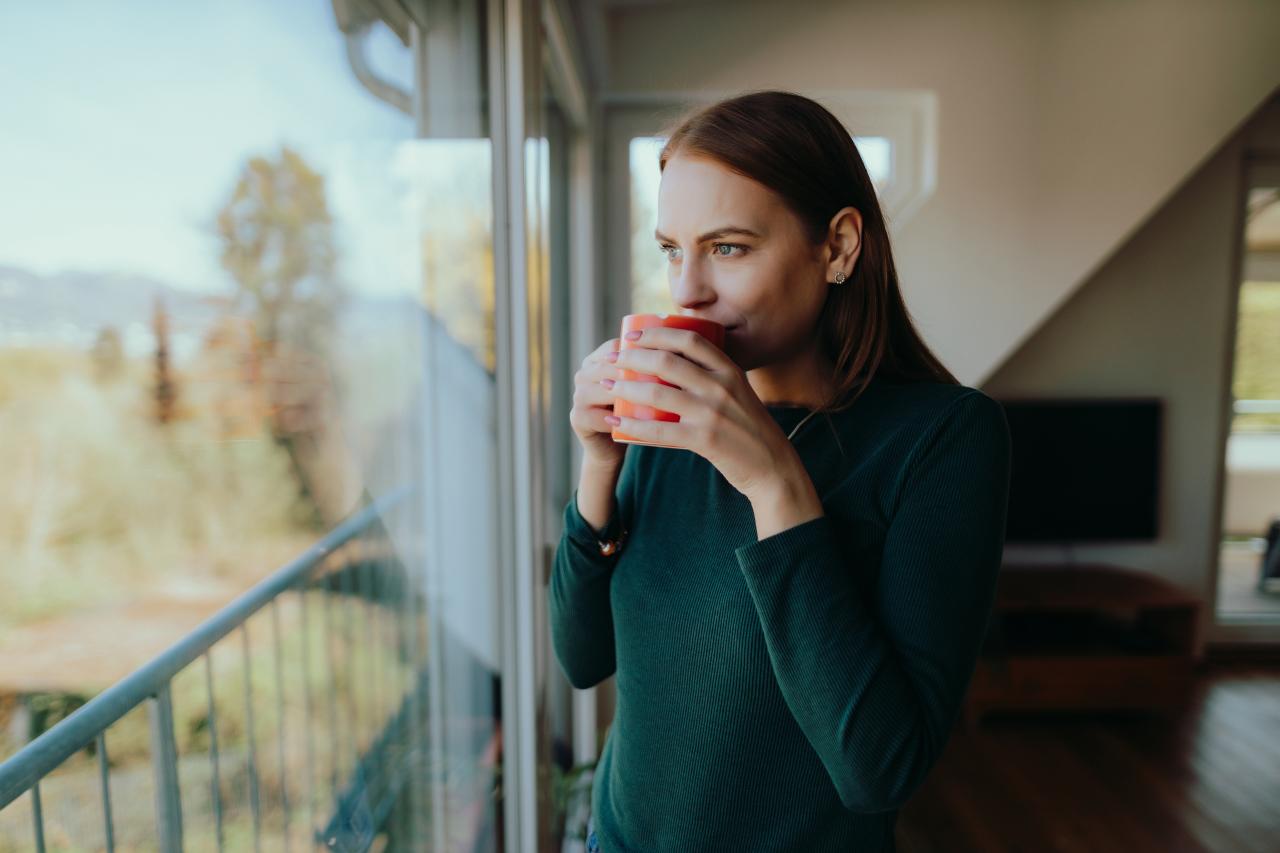 Frau steht an Fenster und nimmt einen Schluck Kaffee